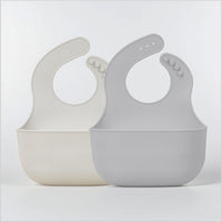 Two YOBIB motion and travel sickness bibs (beige and grey) with deep catch pockets on a light grey background.