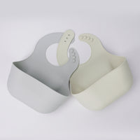 Two YOBIB travel sickness bibs in grey and beige, arranged horizontally, showing deep catch pockets and flexible silicone on a light grey background.
